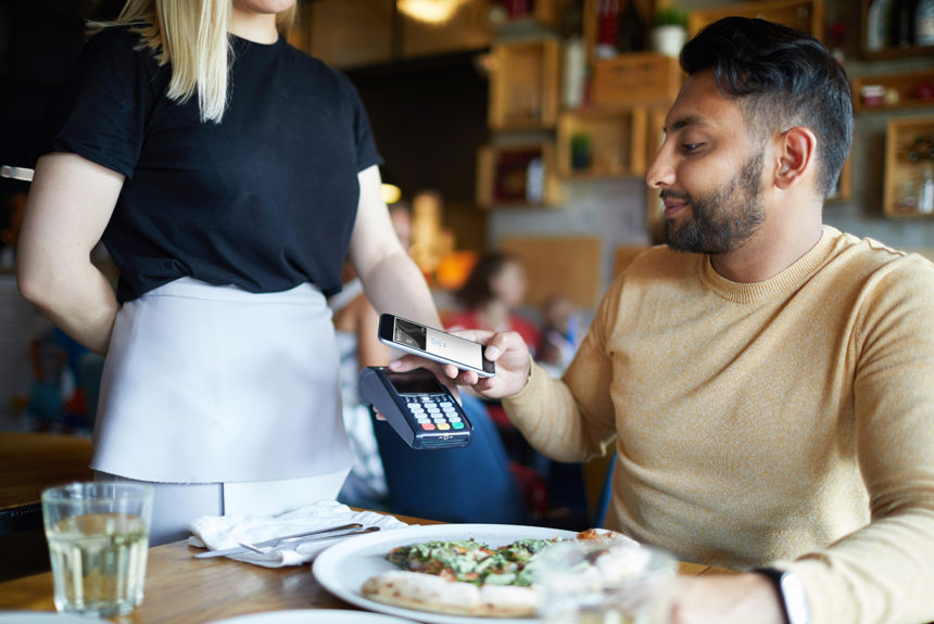 Young man holding his smartphone over payment machine while sitting by table in cafe during lunch