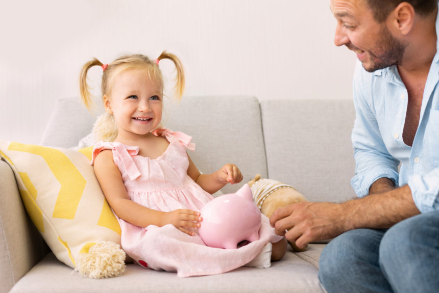 Millennial Father Teaching His Daughter How To Save Money. Cute girl sitting with piggy bank