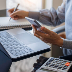 Business woman hand using mobile smartphone and writing on notebook, work on laptop computer with calculator on the desk at modern home office. Online working, work at home concept. Send and Receive Funds Anytime, Anywhere with Our Digital Services